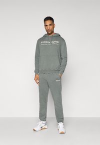 Szürke kapucnis pulóver és melegítőnadrág szett, elülső zsebbel, nyomott logóval és rugalmas mandzsettákkal. Fehér sportcipővel viselve.