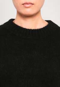 Parte inferiore del viso e collo di una persona che indossa un maglione nero in pelliccia sintetica con scollo a girocollo a coste, su uno sfondo chiaro e semplice.