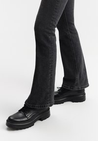 Scarpe in pelle nera con lacci e suola spessa, abbinate a jeans a zampa d'elefante in denim scuro, evidenziando il contrasto di texture e colore.