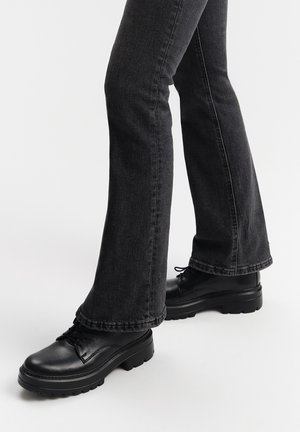 Scarpe in pelle nera con lacci e suola spessa, abbinate a jeans a zampa d'elefante in denim scuro, evidenziando il contrasto di texture e colore.