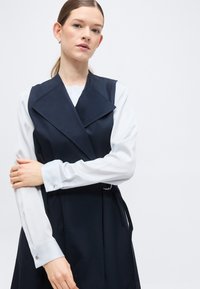 Γυναίκα με ανοιχτόχρωμο δέρμα και καστανά μαλλιά, φορώντας λευκό πουκάμισο με μακριά μανίκια κάτω από αμάνικο μπλε γιλέκο με φαρδύ γιακά.