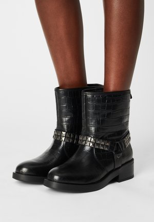 Stivali neri a metà polpaccio con texture a coccodrillo e borchie piramidali argento indossati su gambe nude, in piedi su uno sfondo chiaro uniforme.