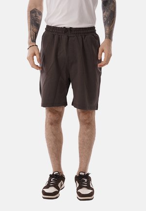 Uomo che indossa pantaloncini marrone scuro a vita elastica, maglietta bianca e sneakers bianche e nere, mostra braccia tatuate su uno sfondo semplice.