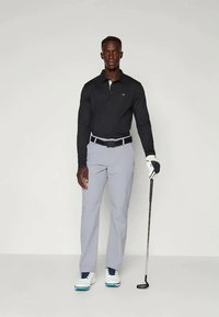 Μαύρο πουκάμισο με μακριά μανίκια, ανοιχτό γκρι παντελόνι γκολφ, μαύρη ζώνη, λευκά παπούτσια με μπλε λεπτομέρειες. Κρατάει μια ράβδο γκολφ, στέκεται σε ουδέτερο φόντο.