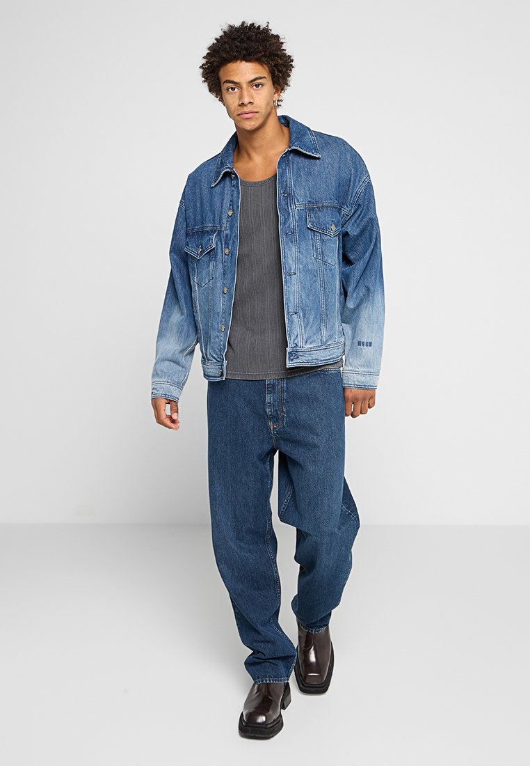 Jeansjacke mit einem steinwaschierten Farbverlauf von hell nach dunkel, ausgestattet mit zwei Brusttaschen. Kombiniert mit einem gerippten grauen Tanktop und lockeren blauen Jeans.