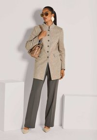 Vrouw in een beige knopenjas, grijze wijde broek, beige schoenen, grote oorbellen, zonnebril en met een bieze handtas met quilt. 