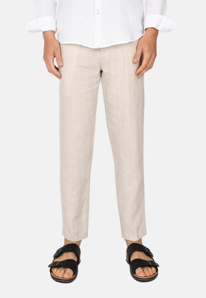 Persona che indossa pantaloni di lino beige a taglio aderente e sandali neri con doppia cinturino, in piedi con le mani rilassate contro uno sfondo semplice.