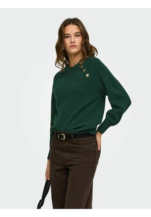 Femme portant un pull vert foncé avec des boutons dorés à l'épaule et un pantalon marron, tenant un sac à main noir avec une main dans la poche.