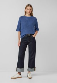 Zils adīts džemperis, brīvi pieguļošs ar īsām piedurknēm, pārī ar tumšiem denim platām biksēm ar salocītiem malām un baltām sporta apaviem.