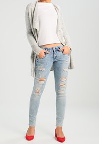 Světle šedý kardigan, bílý strukturovaný top, světle modré roztrhané skinny džíny a červené vysoké podpatky. Kardigan má otevřený přední design.