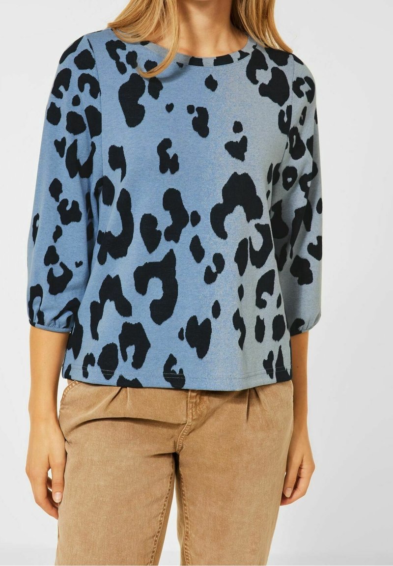 Femme portant un haut bleu à manches longues avec des taches abstraites noires et un pantalon beige, visible des épaules aux hanches.