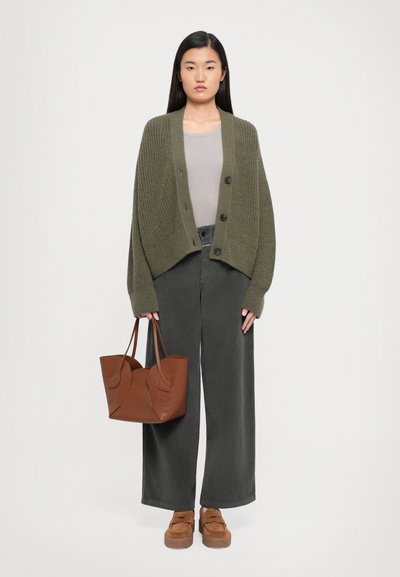 Frau steht und trägt eine olivgrüne Strickjacke, ein hellgraues Oberteil, dunkle weite Hosen und braune Slipper, während sie eine braune Ledertasche hält.