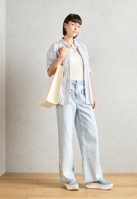 Donna che indossa una camicia a righe blu e bianca con bottoni sopra una maglia beige a costine, jeans a gamba larga in denim chiaro e sneakers blu con zeppa, mentre tiene una borsa a tracolla beige.