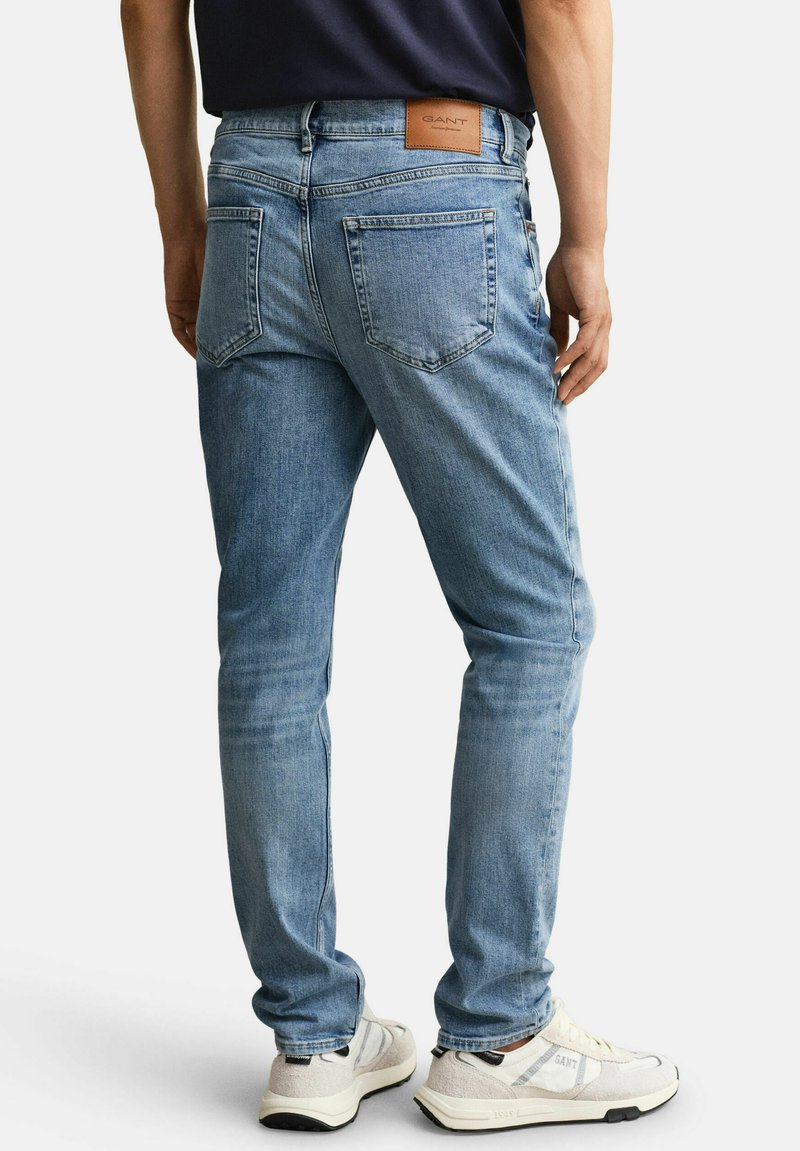 Hombre con vaqueros ajustados azul claro y zapatillas blancas, de pie de espaldas a la cámara contra un fondo liso.