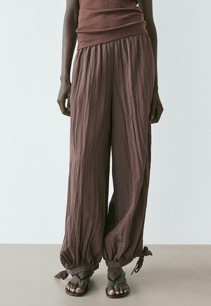 Persona che indossa pantaloni marroni ampi con trama e risvolti raccolti e legati alle caviglie, e sandali neri, in piedi contro uno sfondo chiaro e uniforme.