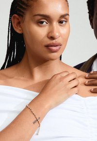 Bracelet en or avec des accents en cristal autour du poignet, porté sur un haut blanc décolleté, avec une texture de peau visible et un tatouage.