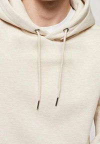 Personne portant un sweat à capuche beige avec cordons, montrant le cou, la poitrine et le haut du torse.