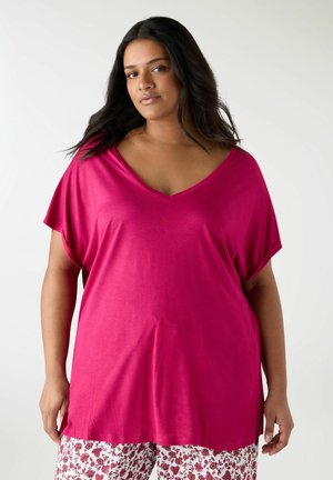 Donna con capelli lunghi e scuri che indossa una maglia rosa acceso a manica corta con scollatura a V e pantaloni bianchi con motivo floreale rosso, in piedi davanti a uno sfondo bianco uniforme.