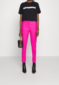 Camiseta negra con el texto "LAGERFELD", combinada con pantalones fucsias ajustados y botas negras de punta. Sosteniendo un pequeño bolso negro.