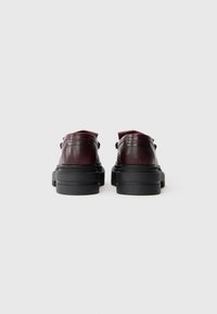 Bordeaux leren schoenen met een gestructureerde afwerking, voorzien van een stevige zwarte zool, ronde neus en veters aan de zijkant. Achteraanzicht.