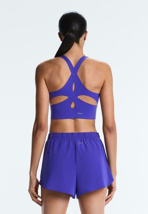 Femme vue de dos portant un soutien-gorge de sport violet à bretelles croisées et un short de sport violet assorti.