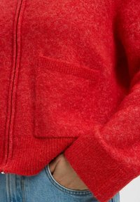 Rode wollen cardigan met een ritssluiting, zichtbare textuur, ribbels aan de mouwen en een zak op de linkerzijde. Casual pasvorm.