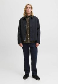 Jonge man met lang blond haar, draagt een zwart jack, een olijfgroen overhemd met knopen, donkere jeans en zwarte schoenen, staande tegen een effen achtergrond.