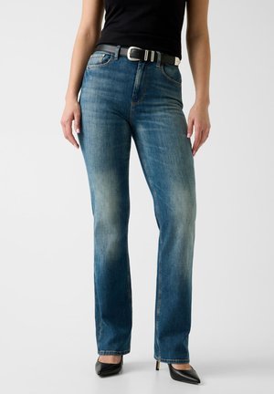 Donna che indossa jeans a gamba dritta blu slavato, cintura nera, top nero senza maniche e décolleté nere con punta appuntita, in piedi davanti a uno sfondo semplice.