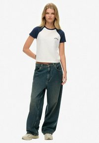 Camiseta raglán blanca y azul marino con el logo "Superdry", combinada con jeans baggy de color azul oscuro. Usada con zapatillas de color claro. Fondo blanco plano.
