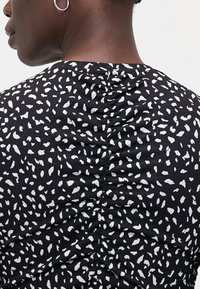 Tissu noir avec des taches blanches irrégulières ; design texturé froncé au dos, col rond et manches courtes. Accents avec un éclat subtil.