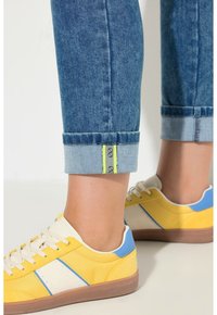 Vaaleansiniset farkut, jotka on käännetty resorista ylös, ja niissä on neonkeltaiset yksityiskohdat sekä beiget tennarit, joissa on keltaisia ja sinisiä paneeleja, valkoiset raidat ja kumipohja.