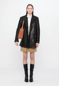 Melnas ādas divrindu žakete, balta krekla, brūna svārku, līdz ceļiem melnas kurpes un brūna pleca soma, parādīta uz neitrāla fona.