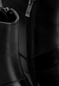 Botines de cuero negro con cremallera lateral, textura lisa y un detalle de panel horizontal. Primer plano que resalta los detalles de la costura.