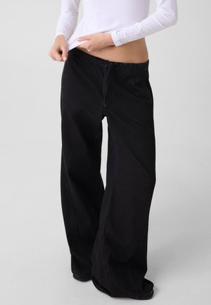Person trägt lockere schwarze Hose und ein weißes Langarmshirt, hält den Hosensaum leicht vom Körper weg.