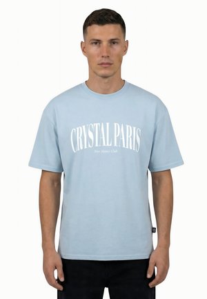 Un homme se tient face à nous, portant un T-shirt bleu clair avec le texte « CRYSTAL PARIS New Money Club » et un pantalon noir sur fond blanc.