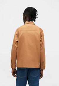 Brun jakke med krage og lange ermer, med knapp mansjetter og sidesplitt. Brukt over blå denim bukser, vist bakfra.