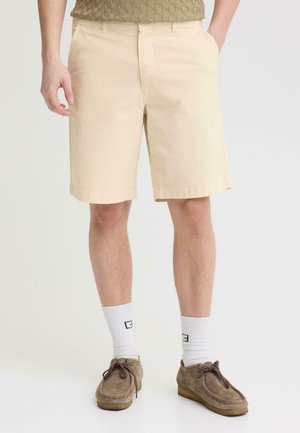 Mann trägt beige knielange Shorts, weiße Sportsocken mit Logo, braune Wildleder-Schnürschuhe, steht mit den Händen in den Taschen.