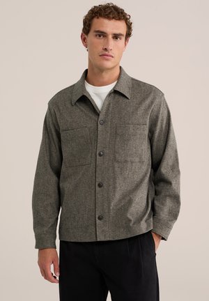Homme aux cheveux bouclés portant une veste-chemise grise boutonnée sur un t-shirt blanc et un pantalon noir, debout avec une main dans la poche.
