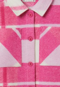 Camicia di flanella a quadri rosa con motivi rosa chiaro e bianchi, due tasche a pattina con bottone e bottoni rosa lungo la parte frontale.