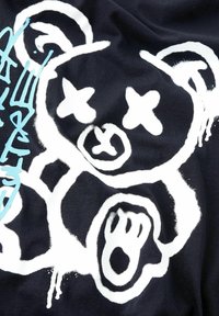 Un tejido negro presenta un gran diseño de oso en estilo graffiti de color blanco, acompañado de formas abstractas. El gráfico está acompañado de texto azul claro.