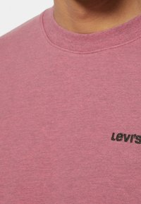Primer plano de una persona que lleva puesta una camiseta de cuello redondo rosa con un pequeño logo negro de Levi's bordado en el pecho.