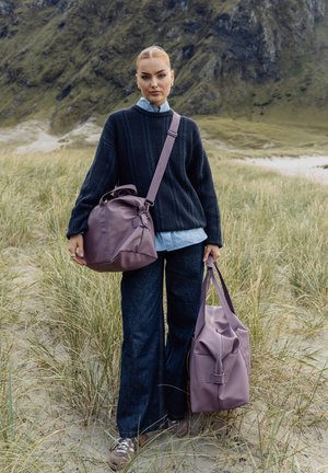 Ung kvinne i svart genser og blå jeans står i høyt gress, holder to lilla vesker, med steinete åser i bakgrunnen.