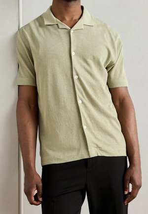 Man in een lichtgroen overhemd met korte mouwen en zwarte broek, staand tegen een eenvoudige lichtgekleurde muur.