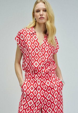 Mujer rubia con camisa de manga corta con patrón geométrico rojo y blanco y pantalones de talle alto a juego, con las manos en los bolsillos.