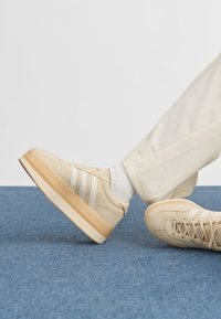 Pessoa usando ténis plataforma bege com riscas brancas, meias brancas e calças cor de creme, com os pés repousando num tapete azul.