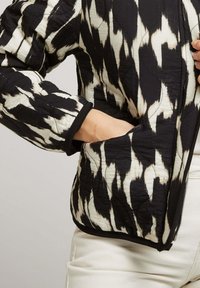 Veste matelassée avec un motif ikat noir et blanc, dotée d'une fermeture éclair à l'avant, de poignets côtelés et de poches latérales avec un éclat texturé.