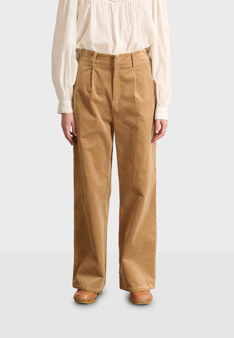 Pantaloni in corduroy di colore marrone chiaro, caratterizzati da una vestibilità ampia, pieghe frontali e una superficie testurizzata. Abbinati a una blusa bianca a maniche lunghe.