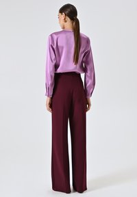 Blusa viola in satin a maniche lunghe abbinata a pantaloni a gamba larga bordeaux a vita alta. Entrambi i capi presentano una texture liscia e un design minimale.