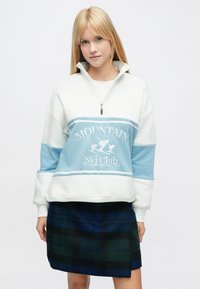 Valge ja helesinine poollukuga pullover, millel on tikitud "Mountain Ski Club", koos tumerohelise ja sinise ruudulise seelikuga.