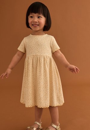 Usmievavé mladé dievča s krátkymi tmavými vlasmi oblečené v svetložltých kvetovaných šatách a zlatých sandáloch stojí s mierne rozpaženými rukami.